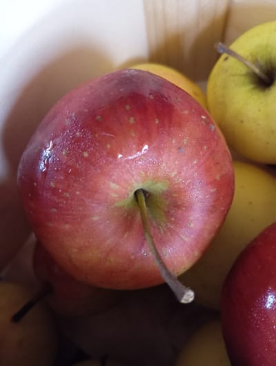 The image is a close-up, top-down view of several apples, with a red apple prominently in the foreground. The red apple is slightly glistening, suggesting it may be wet or polished. It has a stem that is partially visible and extends downwards and to the right. Behind the red apple, yellow apples are visible, one of which has a stem. The apples appear to be in a container, possibly made of wood, indicated by the texture and color in the background. The lighting creates highlights and shadows, giving the scene a warm feel. The location of Hlynytsi, Ukraine, is not depicted or suggested by any visual elements in the image.