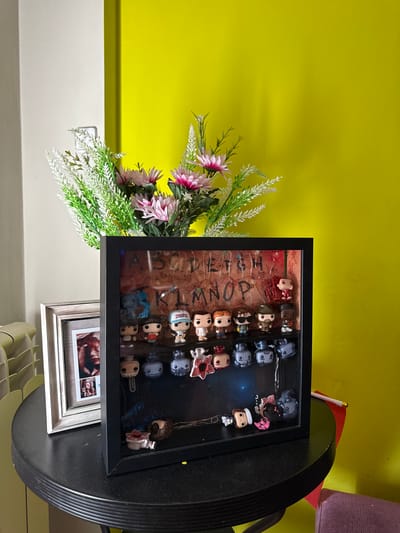 The image shows a framed collection of Funko Pop figurines, likely characters from the TV show "Stranger Things," arranged in a shadow box. The shadow box is placed on a dark, round surface, possibly a table or a turntable, which is itself on a surface that appears to be white. Behind the shadow box, a bright yellow wall is visible, illuminated by what seems to be natural light, suggesting it is daytime. To the left of the shadow box, a bouquet of artificial flowers with pink and white blooms and green foliage adds a touch of color and decoration.
The Funko Pops are displayed in rows within the shadow box. Some of the visible figurines include characters with distinct hairstyles and clothing, characteristic of the "Stranger Things" cast. The background of the shadow box appears to be a collage of images or text, with some letters and lights visible, hinting at the show's aesthetic. One of the figurines, a female character with dark hair, is positioned near the right side of the shadow box. Another figurine, a creature with a flower-like head, is also visible.
Below the shadow box, a smaller framed picture is placed. This framed picture contains what appears to be a close-up image of a person's mouth, possibly a promotional item or a personal keepsake. A small red and gold flag on a stick is also partially visible, emerging from the edge of the round surface. The overall scene suggests a decorative display of merchandise, likely in a home or personal space. The lighting is bright, and the environment seems clean and organized. The location context provided is Getafe, Spain.