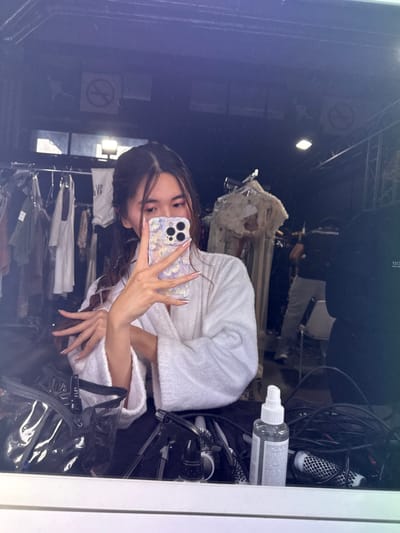 The image depicts a mirror selfie taken indoors within the city of Issy-les-Moulineaux, France. The primary subject is an individual, appearing female, reflected from the waist up, wearing a white terrycloth robe. She holds a light purple, patterned smartphone in her right hand, obscuring the lower half of her face, with her gaze directed towards the phone. Her left hand is raised and extended, with fingers spread, displaying long nails featuring white tips. Her dark hair is styled, exhibiting visible braids or twists.

On the surface immediately in front of the mirror, various items are present: a clear plastic zippered bag with black trim, multiple black hair styling brushes and tools, black electrical cords, and a white spray bottle containing a clear liquid with a partially visible label.

The background, visible in the mirror's reflection, reveals a dimly lit interior space, likely a dressing room or backstage area. Several metal clothing racks are laden with an assortment of garments, including what appears to be formal or theatrical attire, notably a white, possibly embellished garment on a stand to the right. A piece of white fabric or a banner with partial dark text is visible on a rack in the mid-left. Two circular "No Smoking" signs are affixed to the dark-colored wall or ceiling structure. A bright, rectangular light fixture is also visible on the overhead structure. A second individual, appearing male, is partially reflected in the upper right background, wearing a dark t-shirt and light-colored pants, standing and facing away from the mirror.