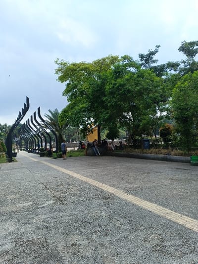 This image depicts an outdoor public space, identified as being in the city of Sarandi de Cuadra, Uruguay, under an overcast sky. The foreground features a light gray, speckled paved walkway that extends into the midground. A yellow tactile paving strip runs diagonally across the walkway from the lower right toward the mid-left.
On the left side of the walkway, a series of approximately ten dark, curved, arch-like structures are arranged in a linear fashion, diminishing in size toward the distant background. Each structure exhibits decorative patterns or script. Potted plants are positioned at the base of some of these structures, and a palm tree is visible behind them. Several individuals are visible walking along the paved path in the distance, partially obscured by these structures.
On the right side of the scene, a raised, dark gray curb separates the paved walkway from an elevated area containing dense green foliage and trees. Two waste bins, one dark blue and one green, are visible within this planted area. Numerous trees with full, green canopies dominate the right midground, with one tree near the center featuring lighter green leaves at its upper sections. Beneath these trees, a group of people is gathered. One male individual, wearing a blue shirt and light-colored shorts, stands facing left, holding a light-colored stick or pole. Adjacent to him, a group of approximately five to seven individuals are seated or lounging on a low wall or bench, partially obscured by foliage, appearing to be socializing. Another individual, dressed in dark pants and a light-colored top, is leaning against a light-colored vertical object or structure, also holding a light-colored stick or pole. The background shows additional trees and obscured structures through the foliage.