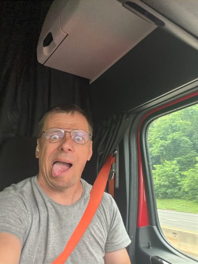A man with glasses is sticking his tongue out and making a goofy face inside a truck cab. He is wearing a gray t-shirt and an orange seatbelt. Through the window, green trees and a road are visible, suggesting the truck is in motion. The interior of the cab is dark with black curtains and a light-colored ceiling. There is no indication of the location being Danbury, United States of America.
