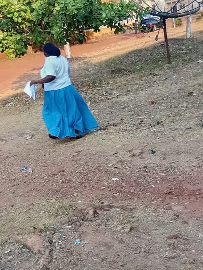 A woman in a light shirt and blue skirt bends over, holding a white paper, on a dry, grassy ground. A satellite dish stands in the background, near a building and a bush. The ground appears to be covered in dry grass and dirt. The scene seems to be outdoors in daylight.