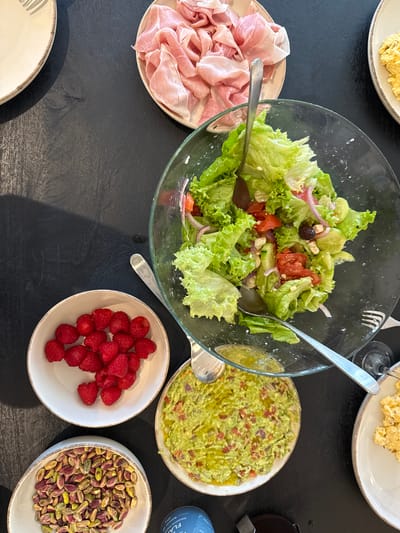 This overhead shot depicts a dark dining table set for a meal in the Municipal Unit of Elliniko, Greece. The table is laden with several dishes and serving items. In the center, a large clear glass bowl contains a fresh green salad composed of lettuce, red onion slices, red tomato chunks, cucumber slices, and black olives, with a silver serving spoon and fork resting within. To the upper right of the salad, a white plate with a brown rim holds a quantity of scrambled yellow eggs. Directly above the salad, another white plate with a brown rim contains thinly sliced pink ham. To the left of the salad, a white bowl with a brown rim is filled with fresh red raspberries. Below the raspberries, another white bowl with a brown rim contains a pile of unshelled green and purple pistachios. Below the main salad bowl, a white plate with a brown rim presents a serving of light green guacamole topped with visible red pieces (likely tomato or onion) and a drizzle of olive oil. Partially visible at the top left is an empty white plate with a brown rim and a fork, suggesting a place setting. A blue container is partially visible at the bottom left. No individuals or direct interactions are visible; the scene presents a prepared spread of food.