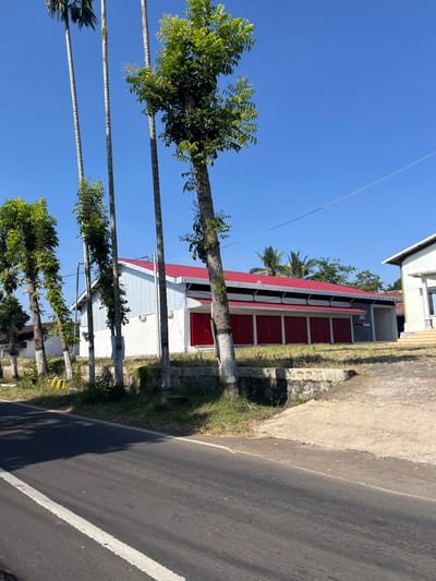 This is a photograph of a building with a red roof and white siding located in Purworejo, Indonesia. The building appears to be a commercial establishment, possibly a warehouse or a shop, with several red doors. In front of the building, there are several tall trees and a paved road. The sky is clear and blue, suggesting a sunny day. The overall scene depicts a typical street view in a rural or semi-urban area of Indonesia.