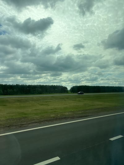 A dark-colored SUV is driving on a paved road. The road has white markings. The vehicle is in the middle of the frame, traveling from left to right. The road is bordered by a green grassy field on the left and a dense line of dark green trees on the right. The sky above is filled with dramatic, overcast clouds with patches of lighter sky visible. The scene appears to be located in a rural or semi-rural area, possibly along a highway in Kirovskoe S/P, Russia.