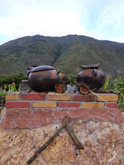 Two dark-colored clay pots rest on a brick structure, which is built upon a larger stone base. The brick structure is composed of red and yellow bricks, while the stone base is textured with shades of brown and white. A broken branch leans against the base of the structure. Behind the pots and brick structure, there is a landscape featuring a mountain range with green vegetation and areas showing signs of burns. Corn stalks are visible on both sides of the structure. The sky is overcast with a mix of grey and blue hues. This scene is located in El Cisne, Ecuador.