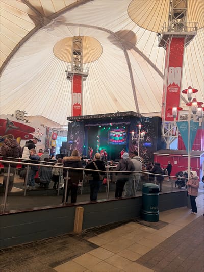 The image depicts the interior of the Skyline Pavilion at Butlin's Skegness resort in East Lindsey, United Kingdom. The scene is dominated by a large, light-colored tensile fabric roof structure supported by internal metal framework and two prominent central pillars adorned with red banners displaying "90 YEARS OF ENTERTAINING THE NATION" and "Butlin's" branding.

At the center-back, a raised stage features an active performance with multiple individuals dressed in red costumes engaged in a choreographed routine. The stage backdrop is illuminated with colorful, geometric patterns and musical note graphics.

A diverse crowd of approximately 20-30 individuals, including adults and children dressed in casual winter attire, are gathered in front of the stage, observing the performance. They occupy a multi-tiered viewing area, separated from the foreground by a metal and glass railing system.

To the left, decorative elements include a large red airplane model mounted on a wall. To the right, several street-style lampposts with multiple red-orange lamps are visible, alongside a red kiosk-like structure. The foreground features a tiled floor in light and dark squares, and a dark green litter bin with "BUTLIN'S" branding is positioned near the railing on the right. The overall environment is brightly lit, primarily by artificial lighting.