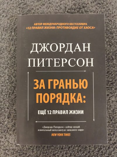 The image shows the cover of a book titled "12 RULES FOR LIFE: AN ANTIDOTE TO CHAOS" by Jordan Peterson. The book is lying on a textured, grey surface, possibly carpet. The cover is predominantly dark grey with white and orange text. The author's name, Jordan Peterson, is prominently displayed in large white font. Above his name, in orange text, it reads "AUTHOR OF INTERNATIONAL BESTSELLER '12 RULES FOR LIFE: AN ANTIDOTE TO CHAOS'". Below Jordan Peterson's name, the main title of the book is presented in large orange font: "ЗА ГРАНЬЮ ПОРЯДКА:". Underneath this, in smaller white text, it says "ЕЩЁ 12 ПРАВИЛ ЖИЗНИ" (meaning "12 MORE RULES FOR LIFE"). At the bottom of the cover, there is a quote in smaller white text attributed to The New York Times, stating "Jordan Peterson - is currently the most influential intellectual in the Western world". There is no indication of the location being in Schwäbisch Gmünd, Germany.