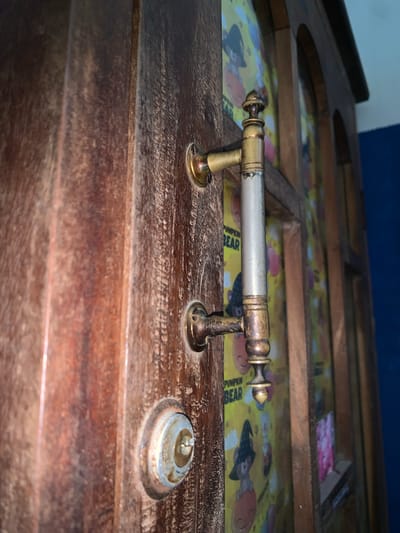 The image shows a close-up, eye-level, side-view of a wooden door with a brass handle and lock. The door has a weathered, textured surface with vertical wood grain. The brass handle is ornate, with decorative ends and a cylindrical central grip that appears to be silver-toned with some wear. Below the handle is a circular brass lock mechanism, showing signs of age and use. Behind the door, a colorful pattern with cartoon bears wearing witch hats is visible, suggesting the door might lead to a themed room or storage area. The background shows the edge of a wooden cabinet or frame and a dark blue wall. The scene is lit from the side, casting shadows and highlighting the textures of the wood and metal. The image does not provide any information about the city of Karangkemiri, Indonesia, nor does it depict any people, actions, or interactions.