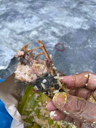 A close-up image depicts a dark-skinned human hand holding a partially consumed piece of food, consisting of fish bones, flaky white meat, dark grey/black skin, and white rice grains. The hand's fingers and palm are stained with yellow and white food residue. Below the held food, more scattered white rice grains are visible on a green banana leaf, which is contained within a crumpled brown paper bag. A blue plastic bag is partially visible on the lower left. The background is a blurry, textured grey surface, likely concrete or asphalt, with a red rubber band visible on it to the right, and a translucent plastic bag on the left. The scene is located in Mns Asan, Indonesia.