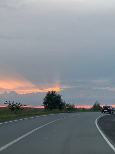 The image shows a road with a car on it, on the side of a grassy field, under a cloudy sky. The sky is dark grey with a band of pink and orange light on the horizon, suggesting it is either sunrise or sunset. The road is grey asphalt with a white line marking the lane. A black car is visible on the right side of the road, moving away from the viewer. There are trees and bushes on the left side of the road and in the distance. The overall scene appears calm and serene. There is no visible text in the image.
