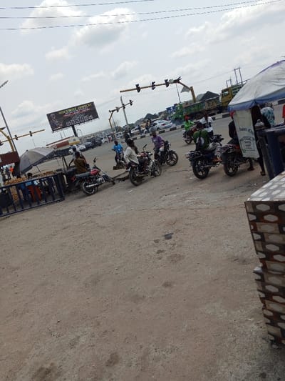 This image depicts a bustling street scene at an intersection in Naze, Nigeria, under an overcast sky. The foreground consists of an unpaved, reddish-brown dirt surface, with a paved road visible in the mid-ground and background. Multiple yellow-painted metal gantries support traffic lights at the intersection.
At least eight motorcycles are visible, many with riders, some appearing to be stopped or moving slowly. A red motorcycle is parked near a street vendor's stall on the left. The paved road features several cars and at least two trucks, including a large green dump truck with a dark load.
On the left side, a street vendor stall with a grey umbrella canopy is set up, accompanied by a blue metal barrier. Another vendor stall with a white canopy is partially visible on the far right, adjacent to a section of a tiled structure in the immediate foreground. A large billboard advertising "GOLDWISE JEWELRY (WORLDWIDE)" is prominent in the mid-ground, above the road. Overhead power lines crisscross the sky. The scene shows multiple individuals engaged in traffic and commercial activities.