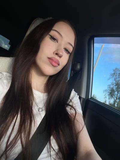A young woman with long, dark hair is taking a selfie in a car. She is wearing a white t-shirt and a silver pendant necklace. The sunroof of the car is open, revealing a blue sky with some clouds and trees in the distance. The scene appears to be set during the daytime. The woman's makeup is visible, with winged eyeliner and light pink lipstick. Her expression is relaxed. The car's interior, including a seatbelt, is also visible. The location is identified as Gavrilovka, Russia.