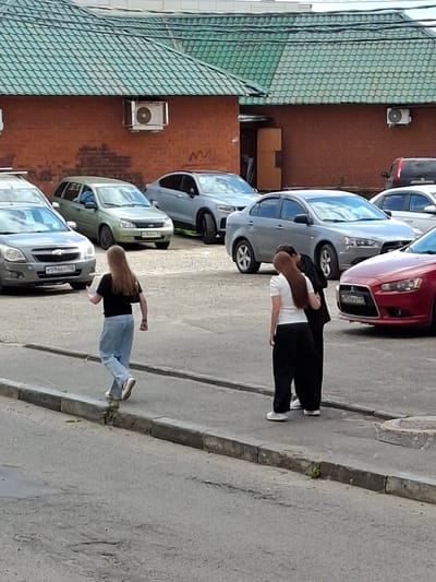 A street-level, medium-shot photograph, taken outdoors on a cloudy day, depicts a scene in Grigorovo, Russia. In the foreground, two individuals stand on a paved area, with a curb and asphalt street in the immediate foreground. The person on the left, viewed from behind, is a young woman with long, light brown hair, wearing a black t-shirt, light blue jeans, and white sneakers. She is walking away from the camera, holding a dark-colored handbag on her left shoulder, and appears to be looking at a smartphone in her right hand. To her right and slightly further back, another individual, possibly a man based on attire, wearing a white t-shirt and black pants, is partially obscured by the first individual. This person is facing away from the camera, and appears to be interacting with a third person, whose back is also to the camera. This third person is wearing a dark jacket over a white t-shirt and dark pants, with dark hair.
In the background, several cars are parked. To the left is a silver sedan, followed by a light green station wagon. Further back, a silver sedan and a red sedan are visible. Behind the parked cars, a brick building with a green, segmented roof is present. Two white air conditioning units are mounted on the brick wall of the building. The environment appears to be an urban or semi-urban area, possibly a parking lot adjacent to a building. The overall lighting is diffused, indicating overcast conditions.