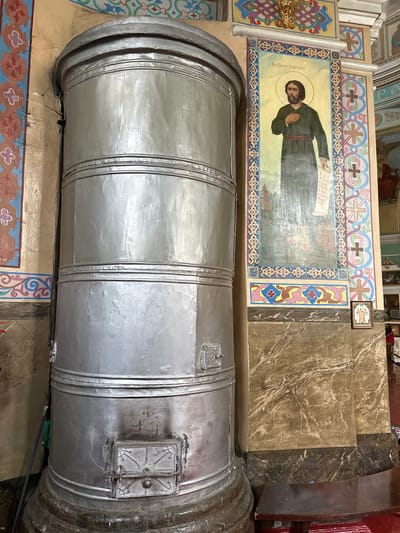 A large, cylindrical, silver-colored stove dominates the left side of the image, positioned next to a column adorned with a religious fresco. The stove has multiple sections, indicated by horizontal bands, and features two metal doors on its front lower section, one above the other. The walls of the building are decorated with colorful, patterned borders and religious artwork. To the right of the stove, a column displays a fresco of a bearded man in dark robes holding a scroll. Below the fresco, a small framed icon of religious figures rests on a marble-patterned surface. A portion of a dark wooden bench is visible in the lower right corner. The environment appears to be an interior space, possibly a church or chapel, with visible ceiling decorations and flooring. The location is identified as Nogornyi, Russia.