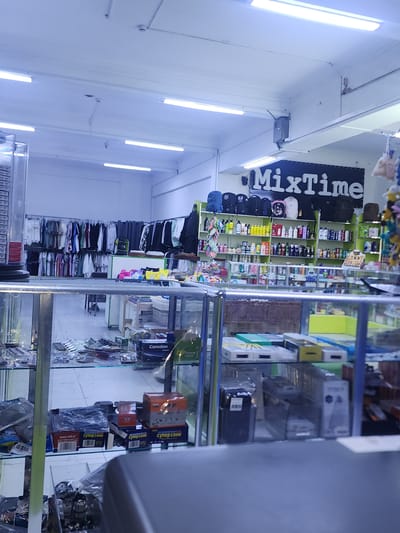 The image shows the interior of a retail store. The store appears to be a general goods store, with shelves stocked with various items including clothing, bags, toiletries, and electronic accessories. The store is well-lit with fluorescent lighting. A sign above the shelves reads "MixTime". The overall impression is of a typical small retail establishment.