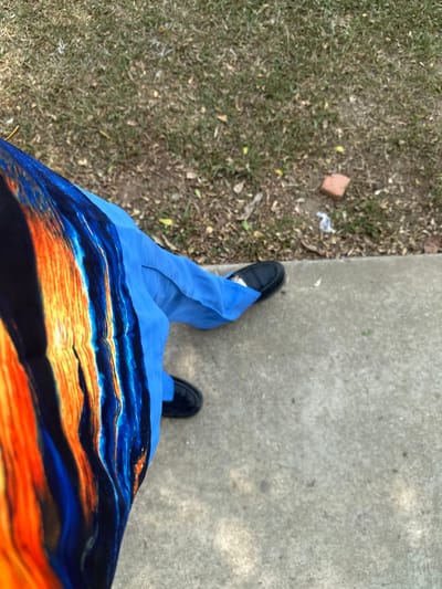 The scene, captured from an overhead perspective in Ozuaha, Nigeria, depicts the lower portion of an individual standing or walking outdoors. The person wears black, flat shoes. Their left leg is partially obscured by a garment featuring a vertical, abstract pattern of black, vibrant orange, and various blue hues. The right leg is clothed in light blue, flowing fabric, likely trousers or a skirt, partially covering the right black shoe. The immediate foreground and right side of the image show a light gray concrete surface. To the left and in the upper background, there is a patch of sparse, dry green grass mixed with exposed brown earth and scattered dead leaves. A reddish-brown brick fragment and a small piece of white debris are visible on the grassy area.