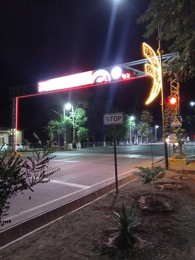 The image shows a street scene at night in Dustlik, Uzbekistan. A large overhead archway displays bright red and white lights. To the right of the arch, a large, illuminated crescent moon decoration is visible, attached to a metal pole. A red traffic light is illuminated on this pole. Below the crescent moon decoration, a pedestrian crossing sign is present. A "STOP" sign is also visible on a pole in the foreground, positioned near a crosswalk marked on the asphalt road. Several trees with visible trunks and foliage are scattered throughout the scene. The ground in the foreground appears to be dirt with some sparse vegetation. Two white cars are parked on the left side of the image, near a building. The overall lighting suggests it is nighttime, with artificial illumination from the arch, decorations, and streetlights.