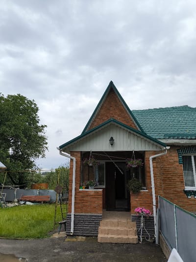 A forensic examination of the image reveals a residential property located in Pysarivka, Ukraine, featuring a single-story brick house with a prominent front porch. The house exterior is constructed of red brick, with a darker, possibly gray or black, stone veneer foundation. The roof is covered with teal-green shingles, and a steep, triangular gable constructed of red brick rises above the front porch. The porch itself is covered, supported by brick columns, and features white vertical siding on its ceiling/frieze, below which a black wall-mounted lantern is affixed. Two white rain downspouts are present on either side of the porch's front columns. The porch entry consists of three light-colored, possibly concrete or stone, steps leading to a dark, open doorway.

Decorative elements include two hanging flower baskets with purple blossoms suspended from the porch ceiling, several potted plants on the porch ledge, and a white metal stand holding pink flowers positioned to the right of the steps. To the right of the house, a low, grey fence or partition is partially visible. The immediate ground in front of the porch is dark, likely concrete or asphalt, with a visible puddle. To the left of the house, a patch of green grass is present, beyond which a dark wooden bench and a metal structure (possibly part of a swing set or other outdoor equipment) are discernible. A large, mature tree with dense green foliage occupies the far left of the frame. The sky is overcast with gray clouds. No individuals are present, nor are any actions or interactions depicted.