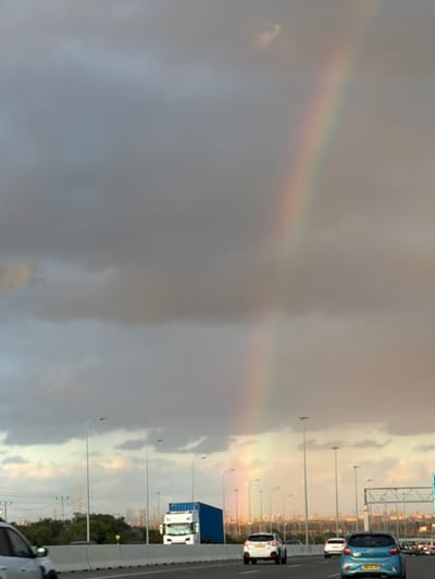 A pale rainbow arcs across a stormy, grey sky beside a highway. Cars and trucks drive on the right side of the frame, illuminated by a hazy, yellowish light. Streetlights line the road. The rainbow is subtle against the heavy cloud cover, with soft bands of color visible. The contrast between the dark sky and the bright highway creates a dramatic scene.