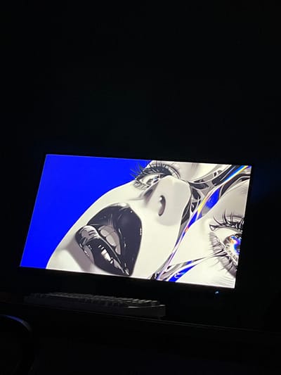 A flat-panel display, positioned at an upward and rightward tilt, is illuminated in a predominantly dark interior space at an unspecified location in Zainsk, Russia. The screen exhibits a digital artwork depicting a partial human face, oriented vertically with the chin angled toward the lower-left corner of the display. The visible features include glossy black lips slightly agape, a nostril, and a prominent eye with long, dark eyelashes. Iridescent, metallic, abstract structures with blue and purple reflections are integrated around the eye and bridge of the nose. The iris of the visible eye also displays a complex, colorful reflective pattern. The artwork's background is a solid, bright blue. Below the illuminated screen, resting on a dark, flat surface, is a light-colored remote control with visible buttons, positioned horizontally and partially illuminated by the screen's light. The environment surrounding the screen and remote control is characterized by near-total darkness.
