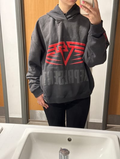 A person is standing in a bathroom. The person is wearing a gray hoodie with red and gray lettering and black pants. The person is holding a smartphone. The background includes a light wood surface and a white sink. The location is assumed to be a bathroom, but the specific city in Ipswich, United Kingdom is not evident from the image.
