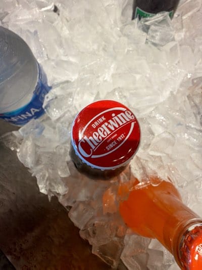 The image shows a close-up of a red-capped Cheerwine soda bottle nestled in crushed ice. Adjacent to it is a bottle of orange-colored soda. Nearby, the upper portion of a water bottle with "AQUAFINA" visible on the label is also partially visible, as well as another dark colored soda in a glass bottle. The scene appears to be taken at a drink station. There are no people visible. The location and other information are not contained within the image.