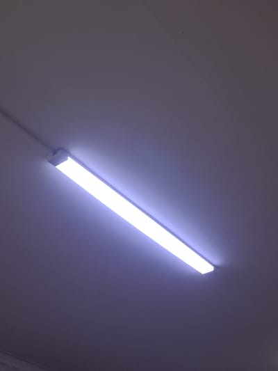 A close-up, angled shot of a white fluorescent light fixture mounted on a gray ceiling. The light is illuminated and casts a pale blue glow downwards. The ceiling appears to be smooth and continuous, with some subtle texture visible. A thin black wire is visible extending from the left end of the light fixture and runs along the ceiling. The scene is devoid of people or any other discernible objects. The lighting suggests an indoor environment, possibly an office or institutional setting. The image was taken in Асықата кенттік әкімдігі, Kazakhstan.