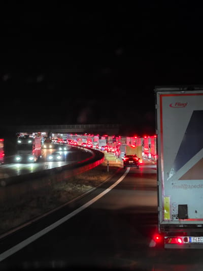 The image shows a traffic jam at night on a highway in Uettingen, Germany. Numerous trucks and cars are stopped, with their brake lights illuminated. The road appears wet, suggesting recent rain. In the foreground, a large truck with "Fliegl" branding is visible, and its rear lights are on. The sky is dark, and the only light sources are the vehicle lights and overhead streetlights. The scene conveys a sense of standstill and congestion.