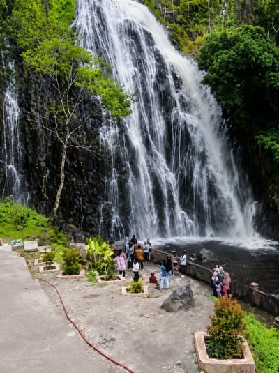 The image shows a wide, multi-tiered waterfall cascading down a dark, rocky cliff face in Sosor Dolok, Indonesia. Lush green vegetation frames the top and sides of the waterfall. In the foreground, a concrete pathway leads to a viewing area where a group of people are gathered. Some are standing and observing the waterfall, while others are interacting in small groups. The people are dressed in casual clothing, with several wearing headscarves. The environment is natural and somewhat developed for tourism, with planters containing small trees and plants along the pathway. The water at the base of the waterfall is dark and appears to be forming a pool.
