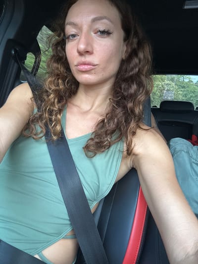 A light-skinned woman with curly brown hair sits in the driver's seat of a car. She is wearing a light green top and a seatbelt. The car's interior is black with a red accent stripe on the seat. Outside the car window, green trees are visible. The location is not possible to determine from the image provided.