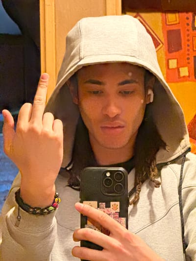 An individual is shown in a close-up, indoor setting within the city of Orsay, France. The subject is a male with a dark complexion and long, braided hair, wearing a light grey hooded sweatshirt with the hood up. A white earbud is visible in his right ear. He holds a black smartphone with a multi-lens camera module and decorative square stickers on its case in his left hand, appearing to be taking a self-portrait. Concurrently, his right hand is raised, with the middle finger prominently extended upwards. Two bracelets adorn his left wrist: one dark-beaded and another composed of multiple brightly colored, spherical elements. The background is out of focus, displaying warm-toned walls; a vertical wooden structure is present on the left, and a wall on the right features abstract geometric patterns in orange, red, and yellow. The lighting is artificial.