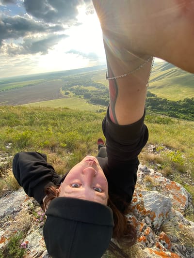 A first-person perspective selfie taken outdoors depicts a young adult with light skin and brown hair, lying on their back on a rocky, grassy slope. The individual is wearing a black beanie, a black hoodie, and dark pants. Their head is tilted back, and their eyes are wide open, looking directly at the camera. A silver nose ring is visible. A tattoo is visible on the left arm, with a silver chain bracelet around the wrist. The background shows a vast landscape with green rolling hills, fields, and trees under a cloudy sky with the sun shining through. The camera is positioned to capture the person from a low angle, emphasizing their position against the expansive scenery. The location is identified as Petrovskoye, Russia.