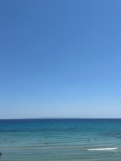This media file displays a seascape with a clear blue sky dominating the left side and the turquoise ocean filling the right. The horizon line is distinctly visible, separating the sky and sea. The ocean is relatively calm with gentle waves and a bright white foamy crest on the far right. Several people are visible in the shallow water, appearing to be swimming or wading. One person is at the edge of the frame on the right, seemingly coming out of the water. The lighting suggests it is daytime, likely a sunny day given the brightness of the sky and the clarity of the water. The scene evokes a sense of leisure and relaxation, typical of a beach or coastal setting. The location is identified as Arkadii Municipal Unit, Greece. No text is visible in the media file.