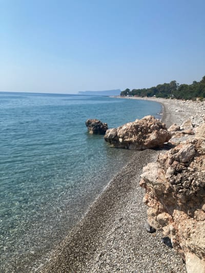 The image shows a pebble beach meeting clear turquoise water under a bright, cloudless blue sky. The shore is lined with numerous grey and white pebbles of varying sizes, with larger, reddish-brown rocks scattered along the waterline and further up the beach. The water is calm and transparent, allowing the pebbles on the seabed to be seen. In the distance, a line of green trees forms the backdrop to the beach, and a faint outline of hills can be seen on the horizon. There are no people or animals visible in the scene. The location is likely a natural beach area in Kumluca, Turkey, given its characteristic rocky coastline and Mediterranean vegetation.