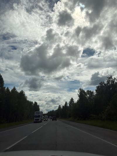 A grey, overcast sky with scattered white clouds dominates the upper portion of the image. The sun is partially visible through a gap in the clouds, casting a bright glow. Below the sky, a dense, dark green forest lines both sides of a two-lane asphalt road. The road stretches into the distance, with several vehicles traveling in the same direction. In the foreground, a white truck is visible, followed by several cars. The scene appears to be a highway or a major road outside of a city or town. The location is identified as Poletaevo 1th, Russia.
