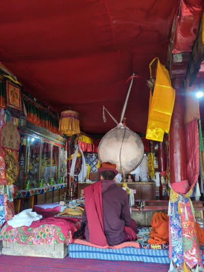 The image depicts the interior of a dimly lit room, possibly a temple or monastery, in Yulang, India. The room is adorned with red fabric draped from the ceiling and walls. In the foreground, a person wearing a maroon robe and a red hat is seated facing away from the camera, meditating or engaged in prayer. They are sitting on a colorful, patterned cushion placed on a raised platform.
The room contains various religious objects and decorations. A large, circular drum hangs from a support structure in the center of the room. Colorful fabrics, including yellow and red streamers, are draped around the space. On the left side of the room, there is a display case or shelf filled with bottles and other items. The overall ambiance suggests a place of worship or spiritual practice, with a rich cultural and religious aesthetic.