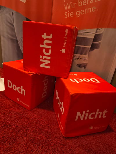 The image depicts three red cube-shaped objects placed on a red carpet. Each cube has white text and a small logo printed on its sides. The text visible includes "Nicht", "Doch", and "Privatkredit". The cubes are stacked and arranged in a way that suggests a display or promotional setup. In the background, a banner with German text is visible. The overall scene appears to be indoors, possibly a promotional event or exhibition. The specific location in Höxter, Germany cannot be determined from the image alone.