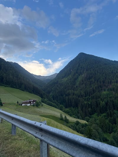 The image captures a scenic mountain landscape in Ratschings - Racines, Italy. The dominant subjects are the lush green mountains covered in dense pine forests and a bright blue sky with scattered white clouds. In the mid-ground, a traditional alpine house with a dark roof and white walls is nestled on a verdant, terraced hillside. The setting is a natural, rural environment, likely during daylight hours given the bright sky and sunlight. A metal guardrail is prominently featured in the foreground, suggesting the vantage point is from a road or overlook. The overall mood is peaceful and serene, highlighting the beauty of the natural landscape. There is no visible text in the image.