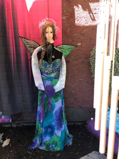 The image shows a mannequin dressed as a fairy. The mannequin has long brown hair adorned with a sparkly headband. It is wearing a shimmering purple and green strapless dress with floral patterns, and sheer sleeves. Green and black fairy wings are attached to its back. The mannequin is holding a bouquet of purple flowers.

The background consists of a pink and gray curtain on the left, and a reddish-brown wooden wall on the right. The ground is covered in dark mulch. To the right of the mannequin, there are tall, light-colored wind chimes and a lush green vine hanging down. In the foreground, there is mulch and some rocks, including a large white rock. A small pink flower petal is visible on the ground near the rock.

The image appears to be taken outdoors, possibly at a market or a garden center. There is no indication of this being in Severn, Canada; this detail is not present in the image itself.