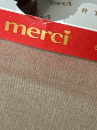 A close-up, angled view in Smyadovo, Bulgaria, reveals a red label with the gold embossed text "merci," where the dot over the 'i' is a small heart shape. To the right of "merci," a faint number "8" is partially visible on the red label. The label's upper edge exhibits slight creasing. Above the red label, fragmented white packaging with faint dark text, including "R T" and "ercl," is visible alongside a blurry dark area, likely the interior of a container or another object. Below the red label, a light brown or beige surface is visible, characterized by a distinct woven diagonal texture. No people are present, and no active interactions are depicted.