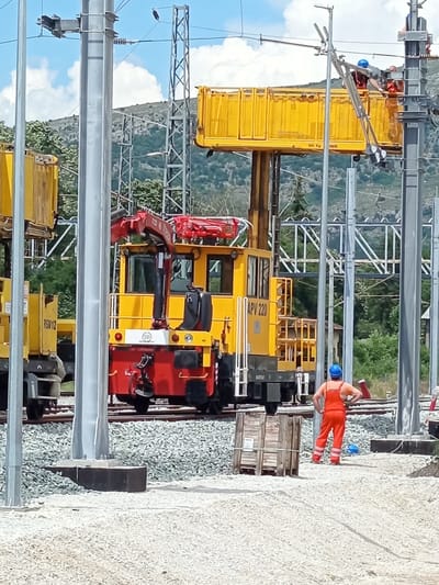 In Dragoman, Bulgaria, on a bright, sunny day with scattered clouds, a construction scene unfolds at a railway line. A yellow and red specialized train with a crane attachment, identified as "APV 220," is positioned on the tracks. Nearby, another yellow train car is visible. A construction worker, dressed in orange overalls and a blue hard hat, stands with their hands on their hips, looking towards the train. In the background, a hillside covered in trees and a structure of metal pylons and wires suggest ongoing infrastructure work, likely related to the railway or electrical grid. The ground is covered in light-colored gravel and some wooden crates are present.