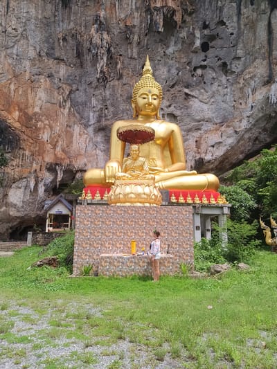 A large, gold-colored Buddha statue sits on a tiled platform in front of a rock cliff. A smaller monk statue is positioned in front of the Buddha, under a red umbrella. A woman stands in front of the platform, facing the statue. The environment is grassy, with trees and a small structure visible in the background. The location is Thap Put, Thailand.