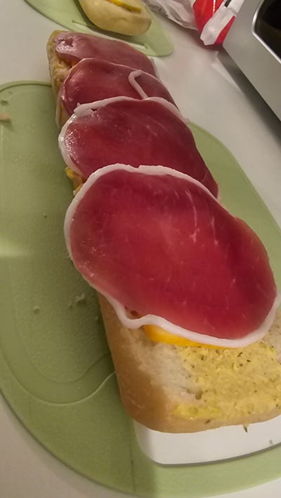 The image depicts a partly assembled sandwich on a light green cutting board. The sandwich features slices of processed ham overlapping each other, placed on top of bread spread with a yellow condiment, potentially mustard or cheese sauce. A partially visible object in the background appears to be a refrigerator or a similar appliance. The setting suggests a domestic kitchen environment. There's no indication of the image being in Rüsselsheim am Main, Germany.