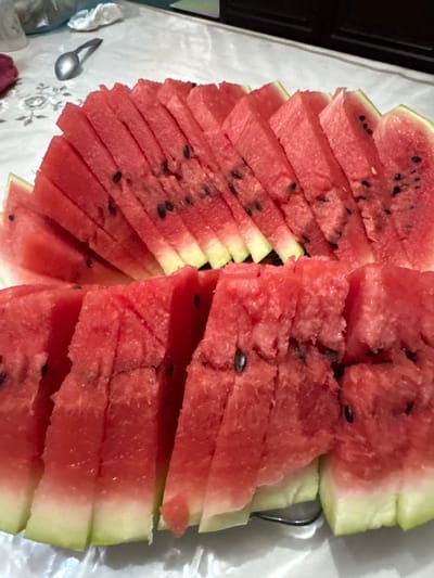 The image shows a platter of sliced watermelon on a white tablecloth. The watermelon slices are arranged in two rows, with the top row slightly elevated and fanned out, and the bottom row forming a more solid base. The watermelon is bright red with black seeds visible throughout. The rind is visible on the bottom of the slices, showing a gradient from green to white. In the background, a silver spoon rests on the tablecloth, near a crumpled white napkin. The tablecloth has a subtle floral pattern. The setting appears to be indoors, on a table. There are no people visible in the image. The location in Batken, Kyrgyzstan, is not discernible from the image itself, as there are no specific geographic markers or architectural details present.