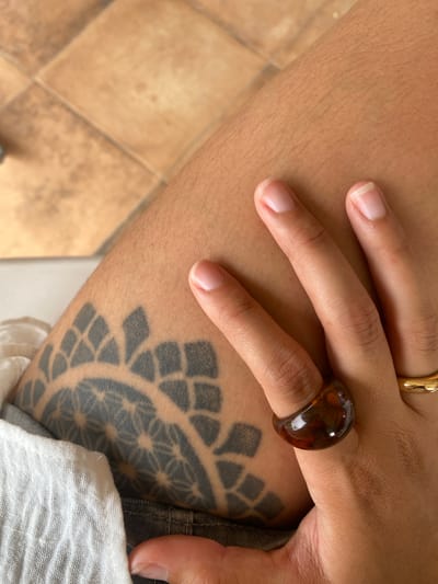 The image shows a close-up view of a person's leg with a tattoo. The tattoo is a black geometric pattern that resembles a mandala or a floral design. The person is wearing a dark ring on their right index finger, and a gold band on their right ring finger. The background consists of light brown tiles. The scene is likely indoors or in a shaded outdoor area, given the soft lighting. The image does not contain any specific landmarks or elements that would allow for identification of its location within Zeneta, Spain.