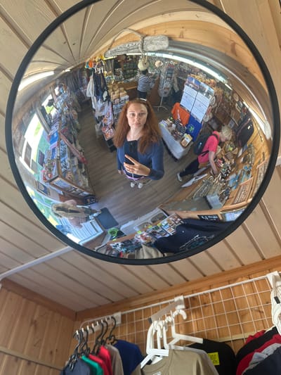 A distorted reflection in a convex mirror captures a woman with red hair taking a selfie. She is wearing a blue long-sleeved shirt and white sneakers, holding a smartphone in her right hand. The mirror reflects a small shop interior filled with shelves of merchandise, including textiles, souvenirs, and calendars. Another woman with blonde hair, wearing a pink shirt and backpack, is visible browsing the shelves in the reflection. The shop appears to be a souvenir store, possibly located in Solovetskiy, Russia, given the context provided. The area outside the mirror shows wooden walls and hanging clothes.