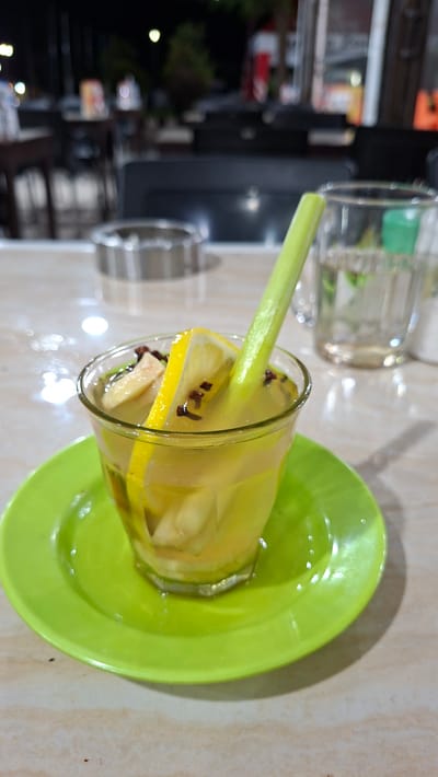 A close-up shot captures a glass of a yellow beverage on a bright green saucer. The drink contains slices of lemon, ginger, and a stalk of lemongrass, which extends out of the glass. The beverage appears to be a refreshing herbal drink. In the background, blurry out-of-focus elements suggest an outdoor seating area, possibly at a cafe or restaurant. The lighting indicates it is nighttime. The table surface is light-colored with a subtle patterned texture. An ashtray is visible in the background, suggesting a smoking-friendly environment. The image provides no specific clues to pinpoint the exact location within Keuniree, Indonesia, beyond it being an establishment with outdoor seating.