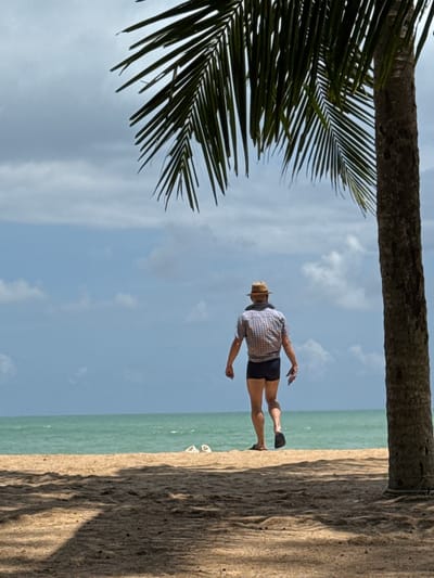 A person wearing a plaid shirt and dark shorts is walking away from the camera on a sandy beach in Ban Khuk Khak, Thailand. The person is also wearing a hat and flip-flops. A palm tree is on the right side of the frame, casting a shadow on the sand. The ocean is visible in the background, with a clear blue sky and some clouds. Two white lounge chairs are visible on the sand in the distance. The scene appears to be a peaceful, tropical setting.