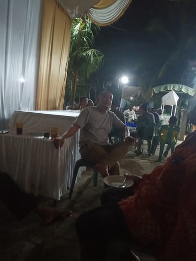 An outdoor gathering is taking place at night, possibly a community event or celebration, in Ulee Ceuruk, Indonesia. The scene is dimly lit by artificial lighting and appears to be in a sandy or dirt area. There are several people present, some seated and some standing, in the foreground and background.
In the foreground, a man in a light-colored t-shirt and khaki pants is seated in a black plastic chair, leaning forward with his arms resting on his legs. He is looking towards the left of the frame. To his right, another person is partially visible, wearing an orange garment with a patterned texture. This person is holding a white bowl with a spoon in it.
In the middle ground, a table draped with a white tablecloth is set up. On the table are at least two glasses, one appearing to contain a dark liquid and the other a lighter colored liquid. A dark rectangular object, possibly a phone, is also on the table. Behind the table, several individuals are seated on plastic chairs. One man in the background, wearing a dark shirt, is looking towards the right. Another individual is seated behind him.
Further in the background, there are palm trees, indicating a tropical or subtropical environment. A white tent-like structure with a green trim is visible on the right side of the frame, and another draped structure, possibly a tent or canopy, is in the center background. The overall atmosphere suggests a casual social gathering.
